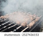 The image shows skewers of marinated meat grilling over hot coals on a barbecue. The meat is sizzling, releasing smoke that rises around the skewers. The skewers have metal handles, and the scene capt