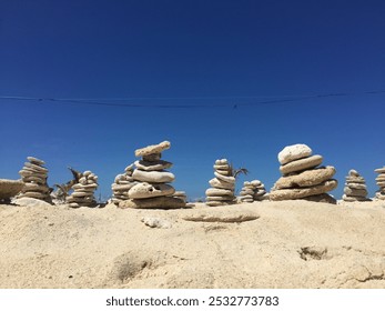 The image shows stacks of smooth stones balanced atop sandy ground under a bright blue sky, creating a calm and minimalist scene. - Powered by Shutterstock