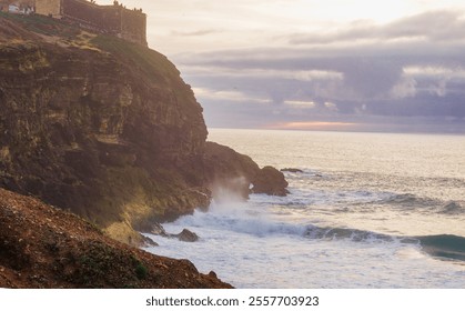 The image shows a stunning coastal scene with waves crashing against a rugged cliffside, topped by a historic fortress. The sky is a mix of clouds and sunlight, creating a dramatic and serene atmosphe - Powered by Shutterstock