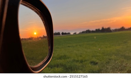 The image shows a sunset viewed through a car’s side mirror, highlighting the contrast between the reflected vibrant sky and the green field in the foreground, creating a serene and thought-provoking  - Powered by Shutterstock