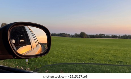 The image shows a sunset viewed through a car’s side mirror, highlighting the contrast between the reflected vibrant sky and the green field in the foreground, creating a serene and thought-provoking  - Powered by Shutterstock