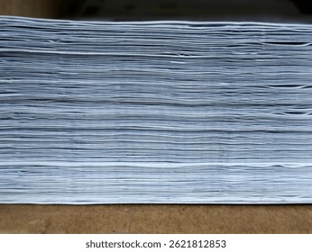 The image shows a thick, uneven stack of white paper viewed from the side, revealing many layers. It rests on a light brown surface.
