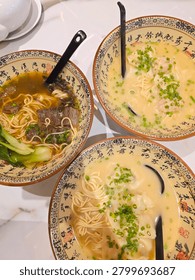 The image shows a top-down view of three large bowls of Chinese noodle soup arranged closely together on a white marble-patterned table.
All three dishes are served in matching, traditional ceramic. The interior of each bowl features an intricate design consisting of black Chinese calligraphic characters and a patterned border around the rim. also includes a long black ceramic soup spoon.
​The Dishes:
​ Contains a dark, rich beef noodle soup. It features thin yellow noodles, chunks of tender braised beef, green bok choy leaves, and a sprinkle of chopped scallions.
​Contains a noodle soup with a light, creamy white broth. It is filled with thin noodles, several wontons or dumplings, and a generous topping of freshly chopped green scallions.
​Bottom Bowl: Similar to the top-right bowl, it contains thin noodles in a milky white broth, accompanied by large wontons or dumplings concentrated in the center and garnished with chopped green scallions.
​Background: In the upper left corner, a portion of a small, white ceramic teapot resting on a matching saucer is visible. The entire setup is illuminated by soft, overhead dining light