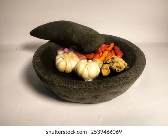 The image shows a traditional stone mortar and pestle filled with various ingredients. Inside the mortar, there are red and orange chili peppers, a bulb of garlic, shallots, and pieces of ginger. - Powered by Shutterstock