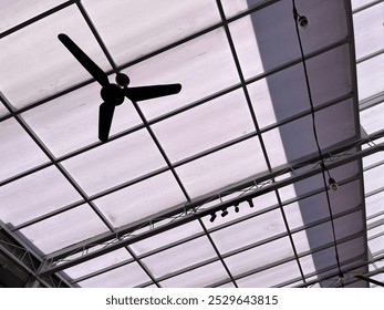 The image shows the underside of a ceiling with a large black ceiling fan in the center, framed by a grid-like structure, with lights and wires hanging down