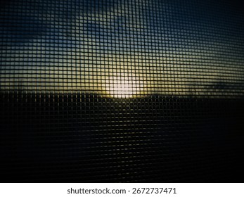 The image shows a view of a sunset through a fine mesh or netting, with the sun partially obscured by clouds. The sky is filled with warm colors, transitioning from deep blue to golden hues.
