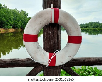 The image shows a white lifebuoy with red stripes hanging on a wooden post near a calm lake. The background features lush green trees reflected in the water, creating a serene atmosphere. - Powered by Shutterstock