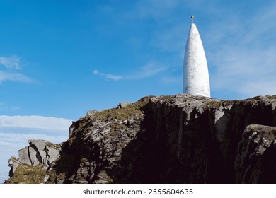 The image shows a white, pointed tower on a cliff edge. The sky above is bright blue with a few wispy clouds.