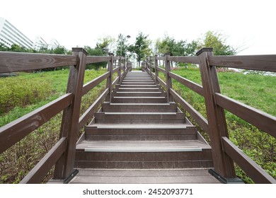 The image shows a wooden staircase with railings leading up a grassy hill. The staircase appears to be part of a walking trail or park, surrounded by greenery and trees. The railings and steps are mad - Powered by Shutterstock