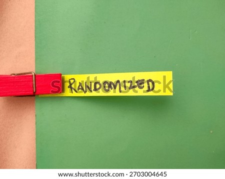 The image shows the word RANDOMIZED written in black marker on a yellow slip of paper, which is held by a red clothespeg against a green background.