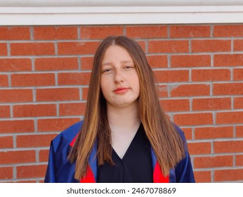 The image shows a young girl with long, straight hair standing in front of a red brick wall. She is wearing a high school graduation outfit. The girl is smiling and looking directly at the camera. - Powered by Shutterstock