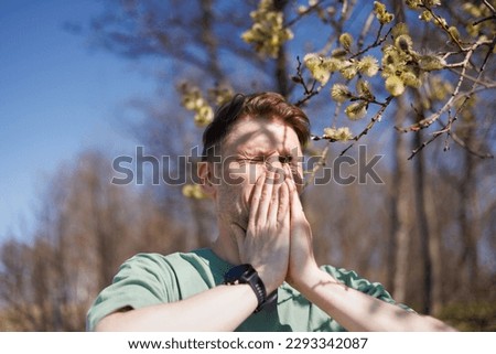 The image shows a young man in a mint-colored shirt sneezing in front of a blooming tree in a park. He appears to be experiencing spring allergies. 