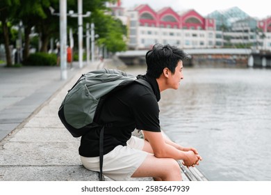The image shows a young man, sitting on a ledge or wall by a river, looking out at the water. He is wearing a backpack and sunglasses