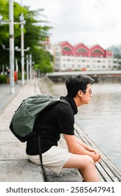 The image shows a young man, sitting on a ledge or wall by a river, looking out at the water. He is wearing a backpack and sunglasses