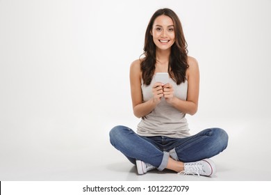 Image of smart woman with long brown hair sitting with legs crossed on the floor using silver smartphone and looking on camera isolated over white wall
