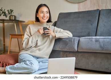 Image of smiling asian woman drinking hot tea, holding cup and sitting near laptop on floor, resting at home. - Powered by Shutterstock