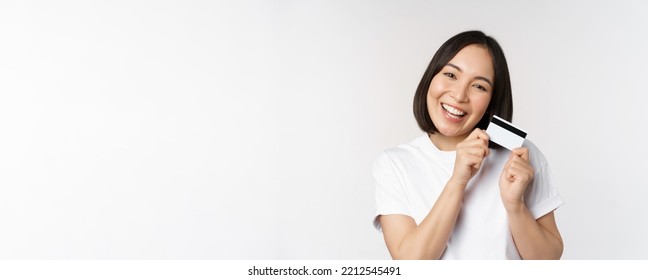 Image Of Smiling Asian Woman Hugging Credit Card, Buying Contactless, Standing In White Tshirt Over White Background