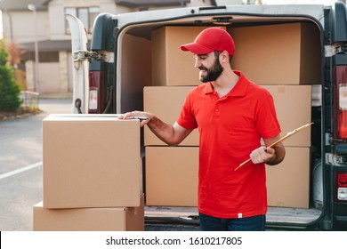 Image Of Smiling Delivery Man In Red Uniform Writing While Standing With Parcel Boxes Near Car Outdoors