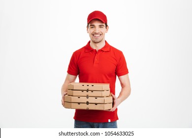 Image of smiling deliveryman in red t-shirt and cap holding stack of pizza boxes isolated over white background