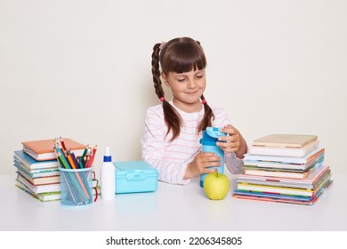 Image of smiling happy little schoolgirl with dark hair and braids sitting at table surrounded with books, holding bottle with water, posing during break isolated over white background. - Powered by Shutterstock