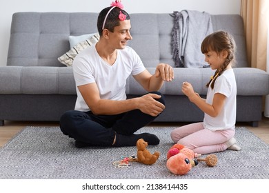 Image Of Smiling Optimistic Man Wearing White T Shirt And Child's Hair Ban With Flower Sitting On Floor And Playing With His Daughter, Cute Female Kid Painting Father Nails.