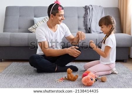 Image of smiling optimistic man wearing white t shirt and child's hair ban with flower sitting on floor and playing with his daughter, cute female kid painting father nails.