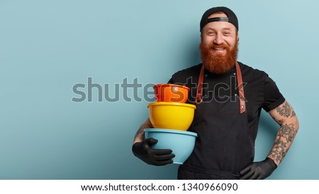 Image of smiling pleased chef holds hand on waist, carries utensils, wears apron and t shirt, being in high spirit for cooking something delicious, stands against blue wall with free space aside