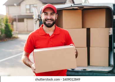 Image of smiling young delivery man in red uniform standing with parcel box near car outdoors - Powered by Shutterstock