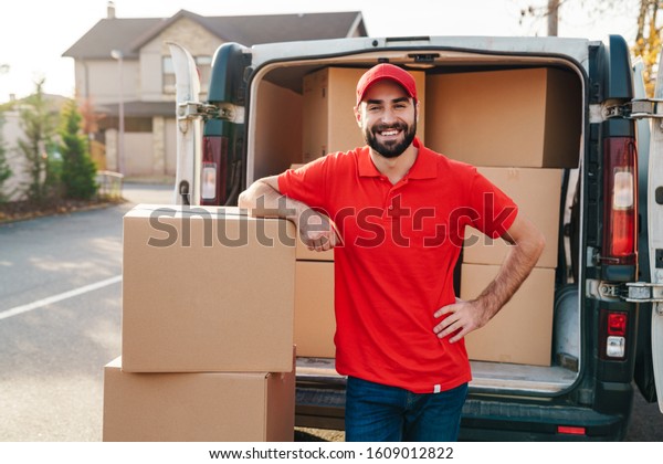 Image of smiling young delivery\
man in red uniform standing with parcel boxes near car\
outdoors