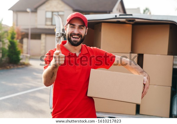 Image of\
smiling young delivery man in red uniform holding parcel box and\
pointing finger at camera near car\
outdoors