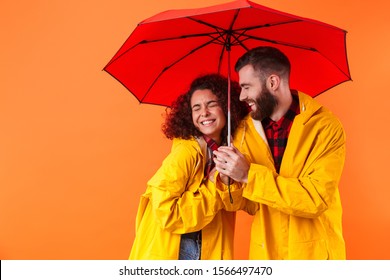 Image of a smiling young loving couple posing in yellow raincoats isolated over orange background with umbrella. - Powered by Shutterstock