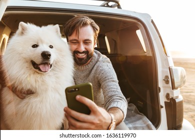 Image Of Smiling Young Man With Dog Samoyed Outdoors At The Beach In Car Using Mobile Phone.