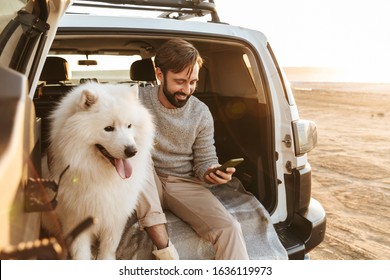 Image Of Smiling Young Man With Dog Samoyed Outdoors At The Beach In Car Using Mobile Phone.