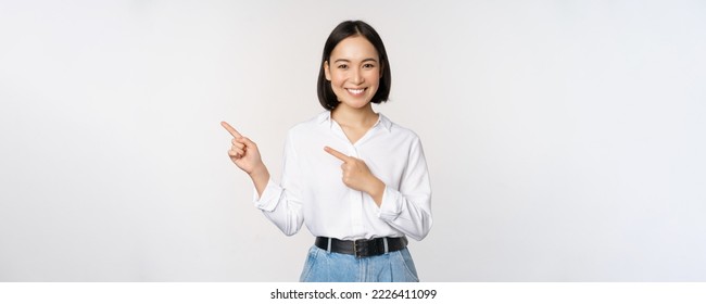 Image of smiling young office lady, asian business entrepreneur pointing fingers left, showing client info, chart of banner aside on copy space, white background. - Shutterstock ID 2226411099