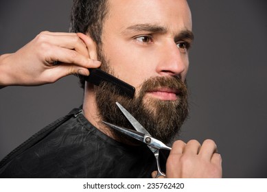 Image as somebody is trimming the beard of a young man on the grey background.
