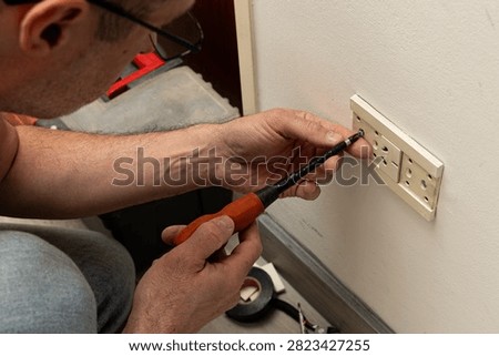 The image is a split view capturing two different moments of an individual working on an electrical outlet in their home. In one section, the person's hands are seen holding a polarity tester over the socket, examining its condition or functionality. This section also shows the individual manipulating small parts within the socket, indicating some form of repair or maintenance activity. The other portion of the image provides a closer look at the actual electrical outlet being worked on. This scene is set against a background that suggests a casual indoor environment.