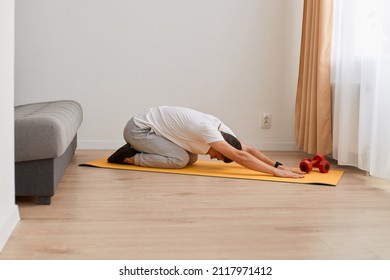 Image Of Sporty Man Stretching And Practicing Yoga At Home, Young Man Doing Exercises, Posing Indoor At Home On Yoga Mat, Training Flexibility, Wearing White T Shirt And Gray Pants.