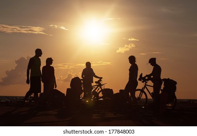 Image of sporty tourist company friends on bicycles outdoors against sunset near the sea and in the clouds. Silhouette people, mountainbike, backpacks, helmets.