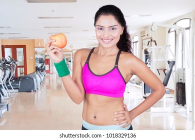 Image of sporty woman smiling at the camera while holding an apple. Shot in the fitness center