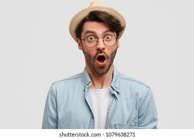 Image of stunned bearded male with surprised expression, wears fashionable hat and denim shirt, being shocked to recieve present from close friend, poses against white background. Fashion and emotions