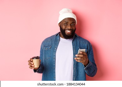 Image of stylish Black man hipster drinking takeaway coffee, reading message on phone and smiling, standing over pink background - Powered by Shutterstock