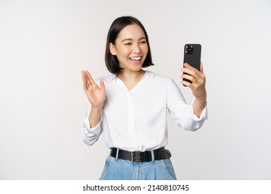 Image of stylish modern asian woman waving hand at smartphone front camera, video chat, chatting with person on mobile phone application, white background