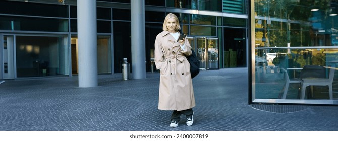 Image of stylish, modern young woman on street, posing in trench coat with backpack, standing and smiling, holding smartphone.