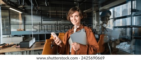 Image of stylish working woman checking her messages on smartphone, holding laptop, standing in an office, working in co-working space.