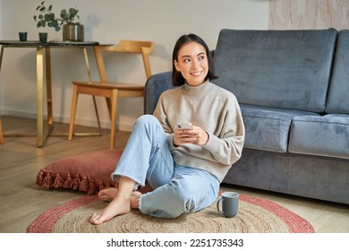 Image of stylish young woman in modern house, using mobile phone, sitting on floor and holding smartphone, drinking from cup. - Powered by Shutterstock