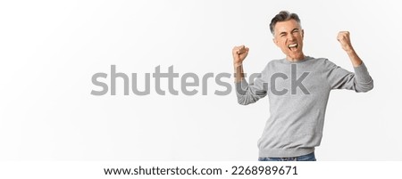 Image of successful middle-aged man triumphing, shouting for joy, raising fists up and celebrating victory, achieve goal, standing over white background.