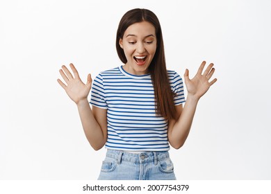 Image of surprised brunette girl smiles, looks in awe down, raise hands and rejoice, found sale promo below, staring at bottomg advertisement, standing against white background