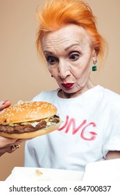 Image Of Surprised Mature Redhead Fashion Woman Standing Isolated At Studio Eating Burger. Looking Aside.