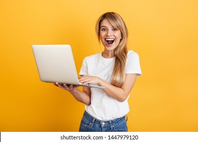 Image of a surprised screaming optimistic emotional young blonde woman posing isolated over yellow wall background dressed in white casual t-shirt using laptop computer.