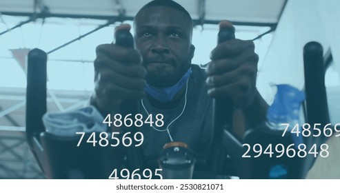 Image of symbols changing over man with face mask exercising on elliptical bike in gym. global covid 19 pandemic, protection and fitness concept digitally generated image. - Powered by Shutterstock